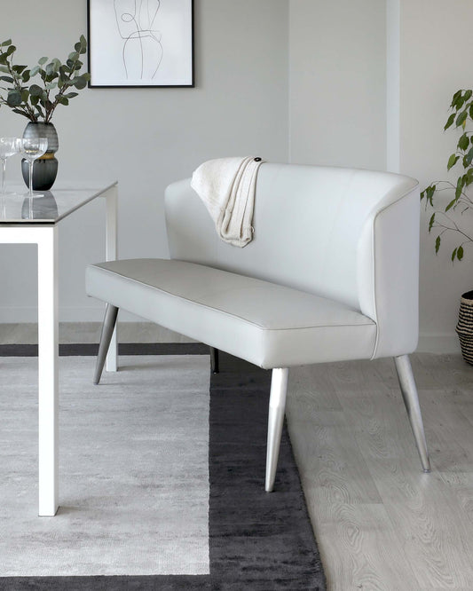 Elegant minimalist-style interior featuring a sleek white leather upholstered bench with a high backrest and angled metal legs, paired with a simple white square side table showcasing a glass vase and small plant. The setting is enhanced by a monochromatic area rug and subtle wall art, creating a modern and sophisticated space.