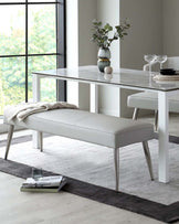 A modern minimalist dining room set, featuring a rectangular white marble-topped table with sleek metallic legs, and a matching bench with a light grey upholstered cushion and simple metallic leg design. The set is showcased in a bright room with large windows and a subtle grey area rug beneath.