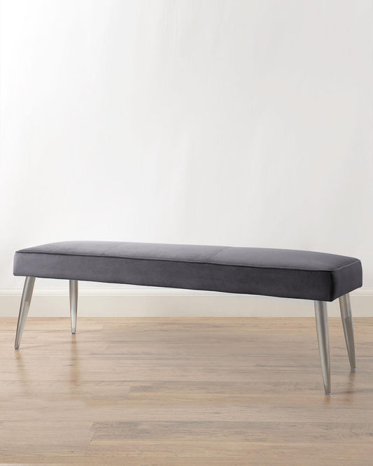 Modern upholstered bench with sleek, charcoal grey fabric and slender, tapered metal legs in a brushed finish, positioned against a neutral wall and hardwood floor.