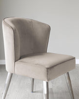 Elegant modern dining chair with a high curved backrest and plush seat upholstered in light taupe fabric, featuring clean lines and sleek, tapered metal legs in a brushed finish.