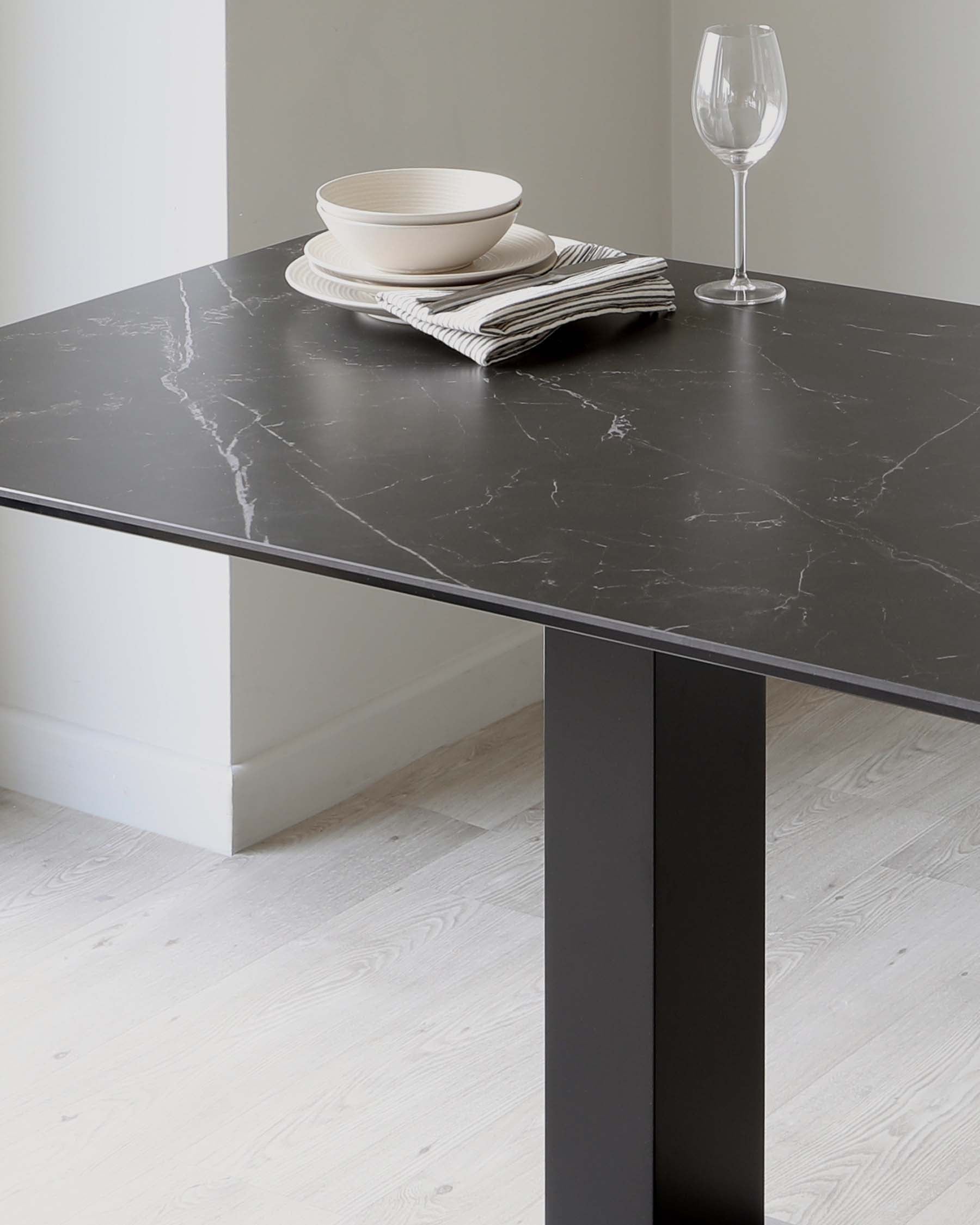 Elegant rectangular dining table with a dark faux marble tabletop and a minimalistic black square leg, set against a light wood floor and pale wall backdrop, adorned with a simple white bowl and plate on striped cloth napkins and a single clear wine glass.