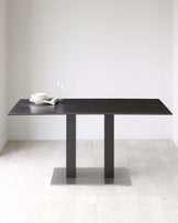 Modern rectangular dining table with black marble top and minimalist dual pedestal base in a matte black finish, displayed on a light wood floor against an off-white wall. Table is partially set with white dishes, a clear wine glass, and silver cutlery.