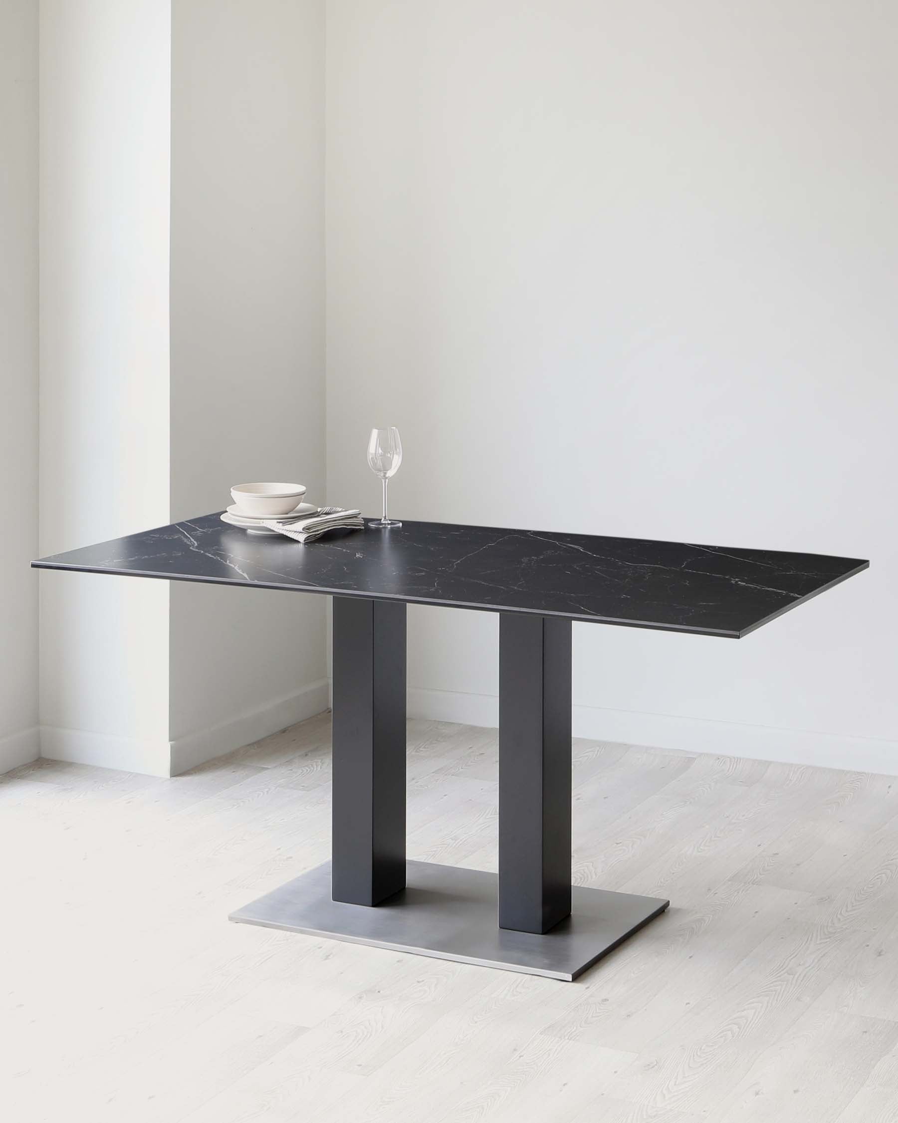 A modern dining table with a black marble top and thick rectangular legs in a matte black finish, set on a minimalist stainless steel base. The table is adorned with a simple table setting including white plates, a napkin, and a clear wine glass.