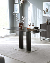 Modern minimalist dining room with a sleek rectangular dining table featuring a dark grey tabletop on bold rectangular pedestal legs with metallic accents, set on a textured off-white area rug. A matching sideboard with vertical groove detailing stands against the wall, adorned with decorative vases and artwork. The room is bathed in natural light with a serene colour palette.