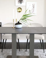 Modern minimalist dining furniture featuring a rectangular table with a dark grey stone-like top and sleek metallic legs, paired with two light beige upholstered chairs with dark angular wooden legs. The table is adorned with a round black vase holding greenery and two tall white candles in gold holders.