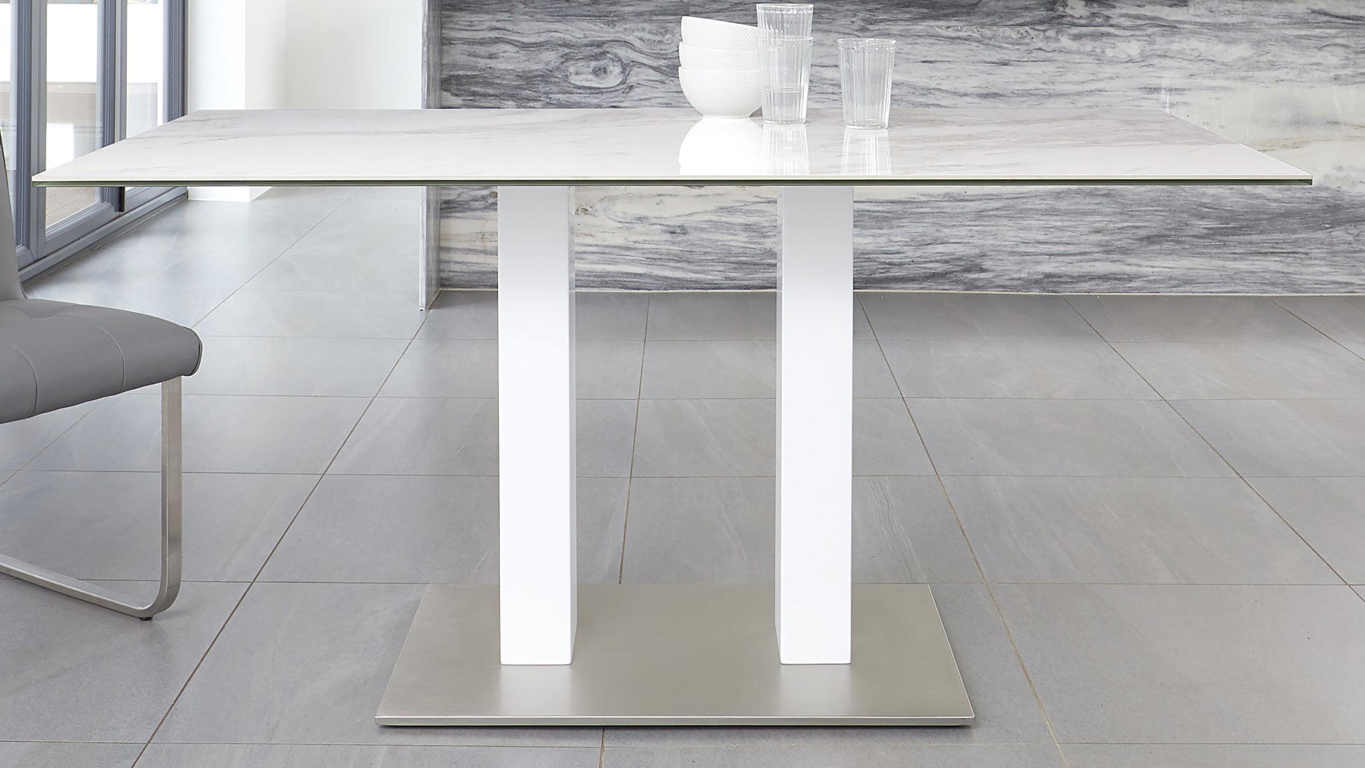 Modern minimalist rectangular dining table with a sleek white marble top and sturdy metallic white legs connected to a rectangular base. The table is adorned with simple, clear glassware.