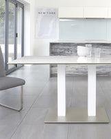 Modern minimalist dining room featuring a sleek, rectangular white dining table with a marble-like surface and sturdy broad legs, paired with a contemporary grey leather chair with a unique metal base. The room is accentuated by a monochrome New York map poster and a set of simple, transparent glassware on the countertop, all against a backdrop of clean white cabinetry and soft grey floor tiles.