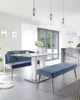 Modern dining room set featuring a rectangular white marble table with a dual pedestal base and contrasting sleek metal support bars. Accompanying the table is a blue velvet upholstered corner bench with backrest and silver metal legs, and a matching blue velvet ottoman bench without a backrest. The set is complemented by minimalist decor and natural light.