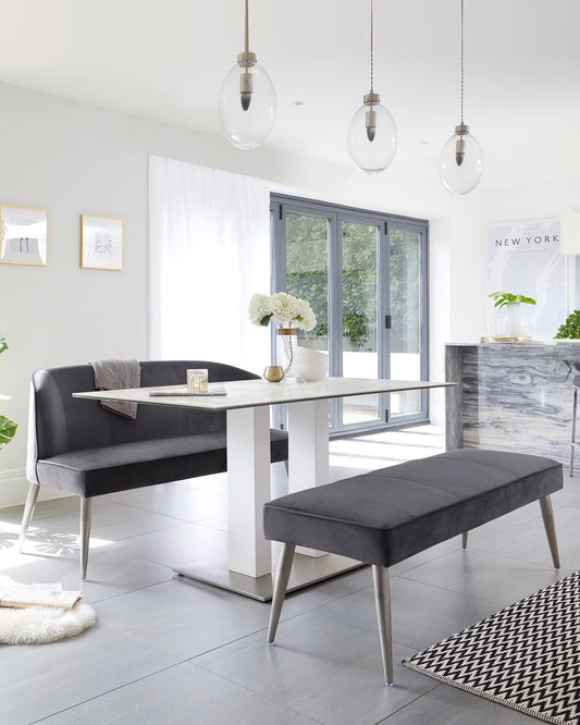 Modern dining room furniture featuring a rectangular white dining table with a glossy finish and sleek metallic accents, paired with a grey upholstered L-shaped dining bench and a matching rectangular ottoman-style bench with metal legs.