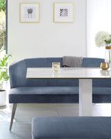 Elegant dining nook featuring a blue velvet upholstered banquette with a smooth curved back, accompanied by a sleek white rectangular table with a reflective surface and a solid base. A matching blue velvet bench sits opposite the banquette, enhancing the modern aesthetic of the set. The scene is accessorized with minimalist decor, including framed artwork, a grey throw, and a golden vase with white flowers, all contributing to a chic and contemporary dining space.