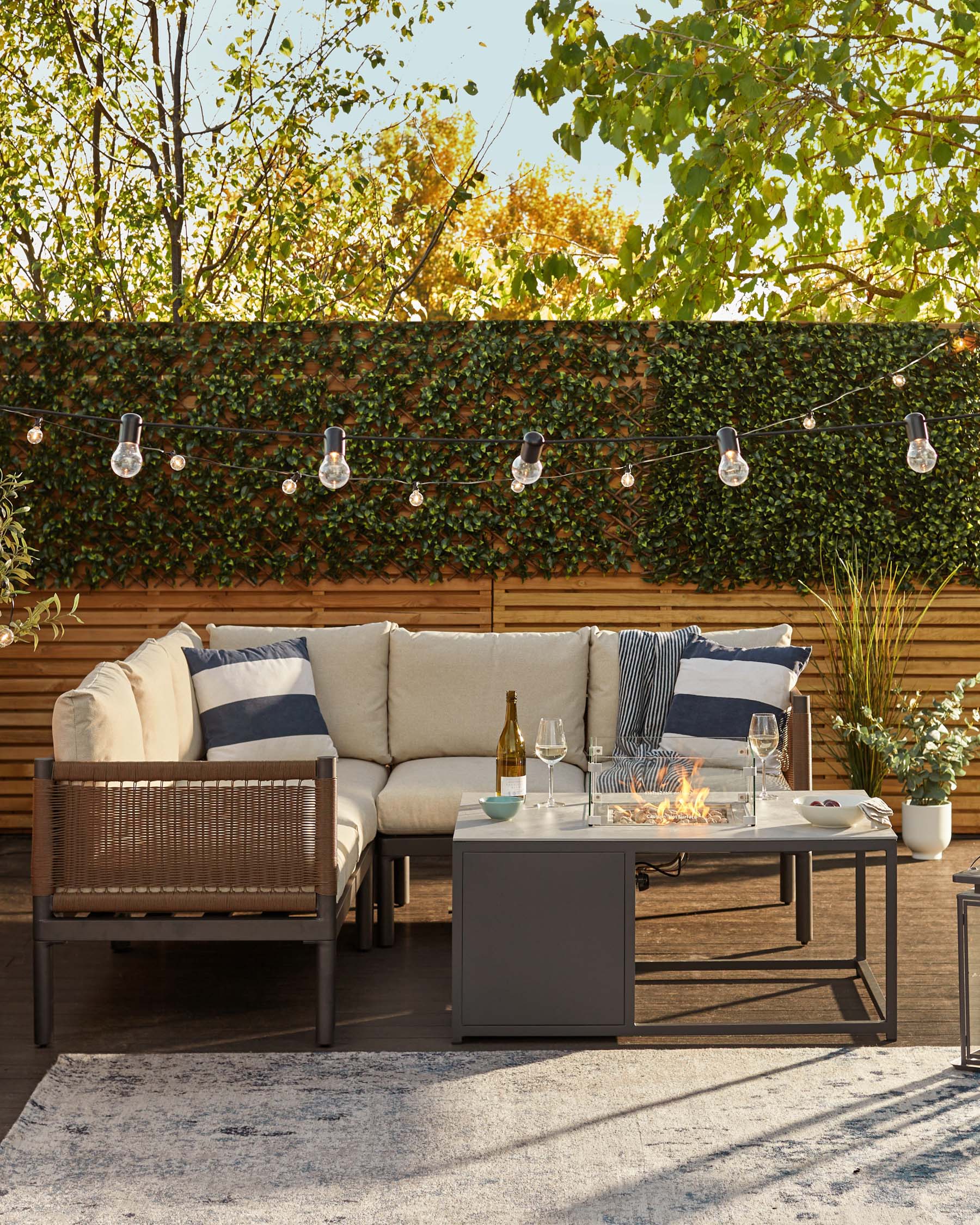 Outdoor furniture set featuring a sectional sofa with cream cushions and dark wood framing, accompanied by multi-striped throw pillows. A matching wood and metal coffee table with a built-in fire pit at the centre, and a small, square, side table completes the set. The arrangement is on a textured outdoor rug, creating an inviting and cosy patio space.