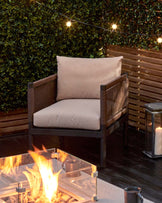 Outdoor lounge chair with a modern design, featuring a sturdy dark frame with wicker accents and thick, comfortable beige cushions, set against an inviting patio backdrop with ambient string lights and a glowing fire pit table.