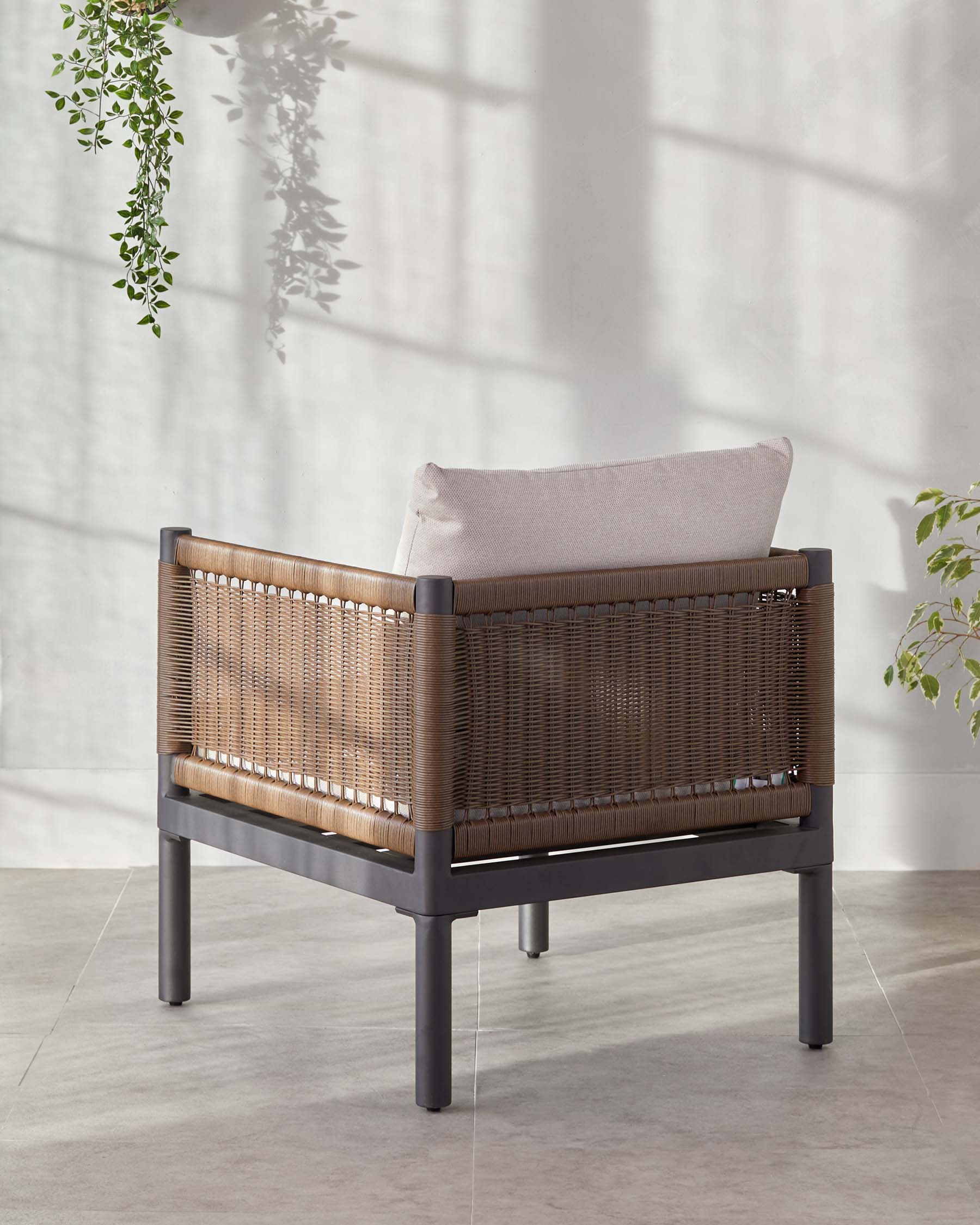 Modern armchair with a dark grey aluminium frame and brown synthetic wicker back and sides, complete with a light beige seat cushion and lumbar pillow. Positioned on a concrete floor with a potted hanging plant casting shadows on the plain wall behind it.