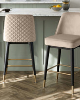 A set of two contemporary bar stools with elegant quilted beige upholstery. Each stool features a high backrest with diamond stitching, a comfortable padded seat, and sleek black legs with gold footrests and gold-tipped feet. The stools are positioned near a kitchen counter with a marble top.