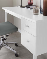 A modern white desk with clean lines, featuring an uncluttered tabletop and two drawers with simple rectangular silver handles. Paired with the desk is a grey upholstered office chair with a metallic base and caster wheels for mobility.