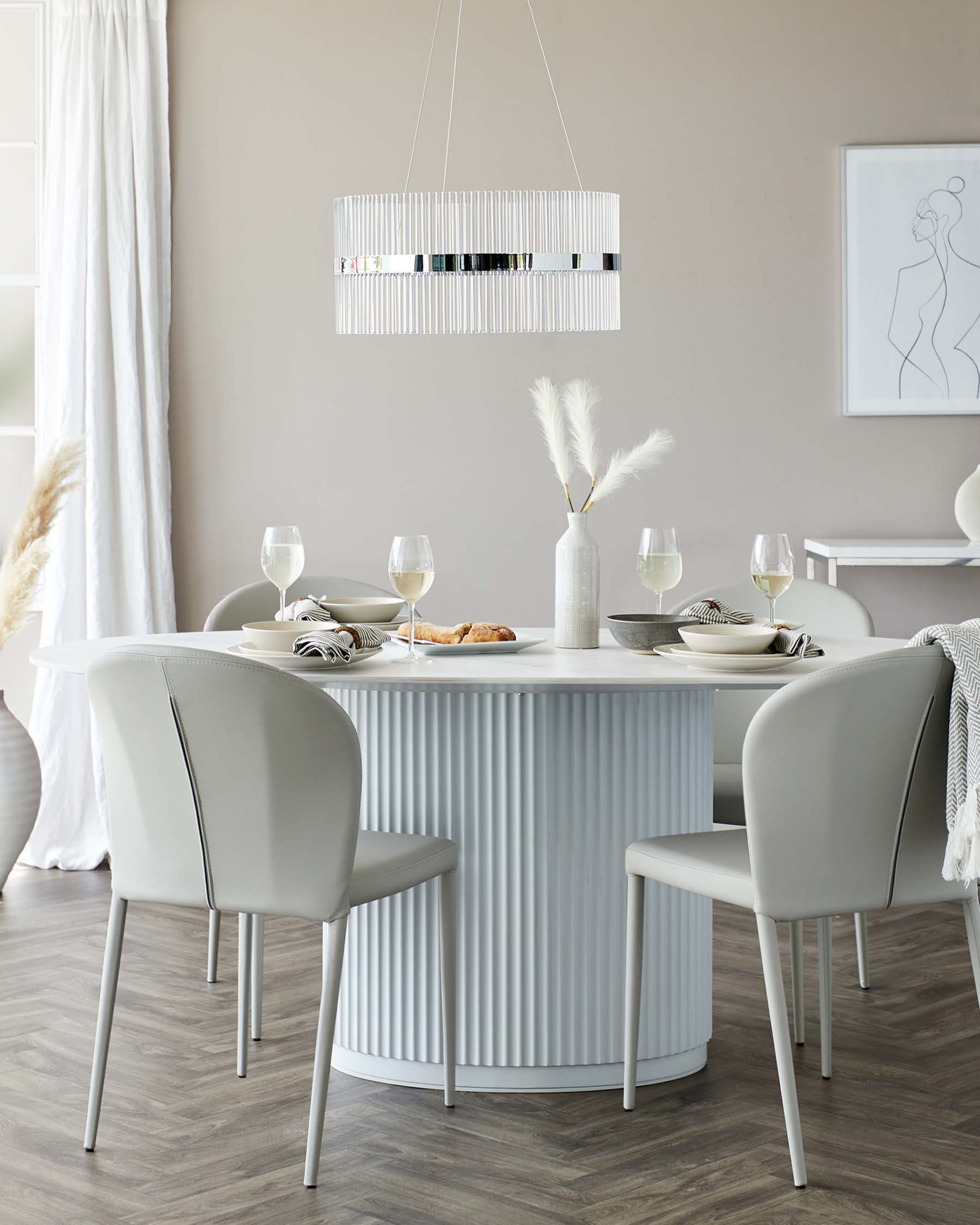 Elegant dining room setup featuring a round, white, fluted pedestal table paired with four sleek, light grey upholstered chairs with slim metal legs. A modern white fringed chandelier hangs above the table. The table is set with minimalistic dinnerware, glasses, and a decorative vase with pampas grass.