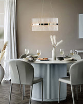 Modern dining room setup featuring a round, ribbed base table with a smooth tabletop, and four sleek, upholstered dining chairs with metal legs. The setting is complemented by an elegant, elongated pendant light with vertical stripes above the table.