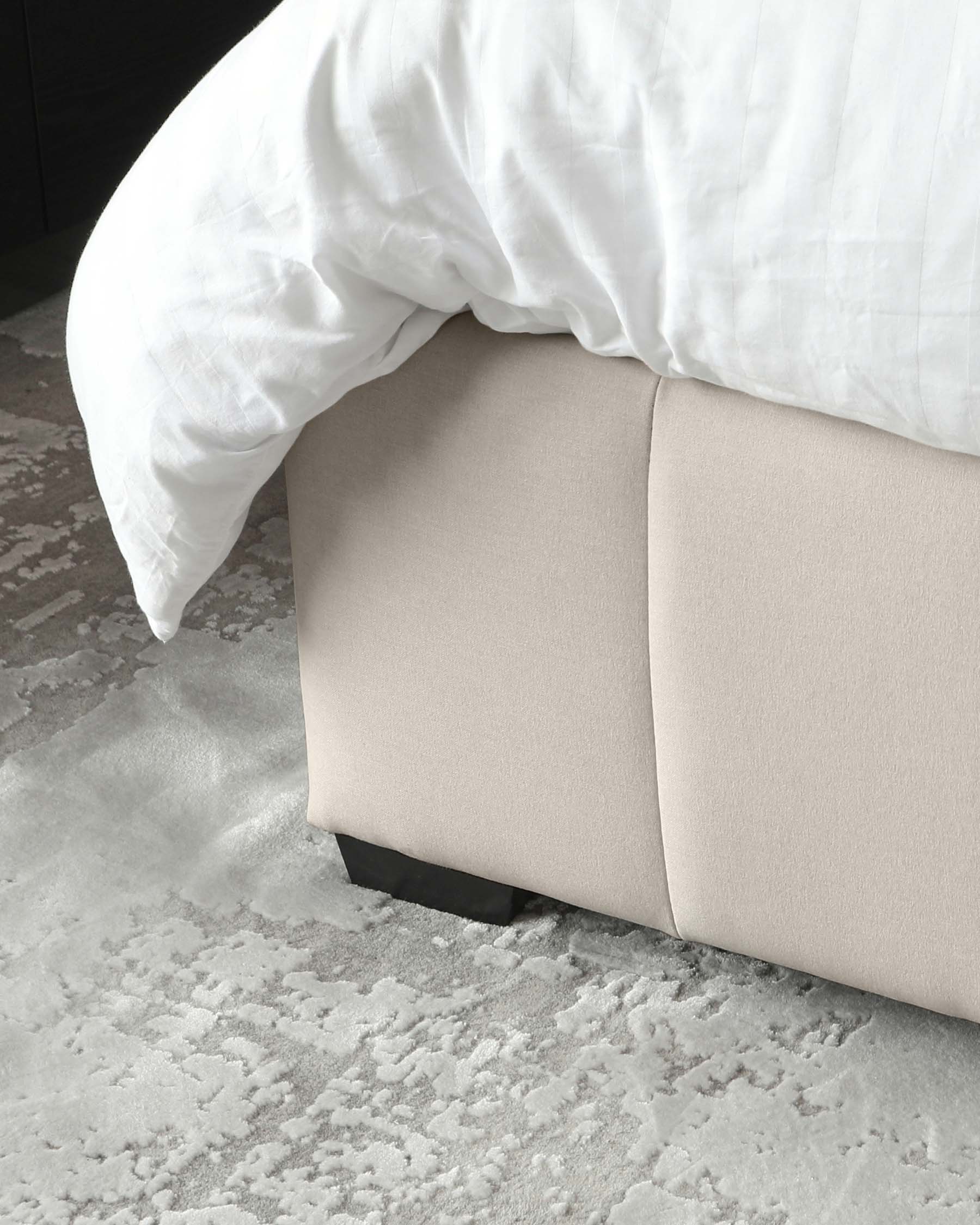 Contemporary bed frame with an upholstered, beige headboard extending to a matching platform base, accentuated with discrete black feet, partially obscured by a draped white duvet on a textured grey area rug.