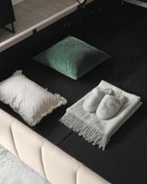 Elegant modern furniture featuring a minimalist beige sofa with a streamlined design. On a dark grey rug lies a plush green velvet pillow, an off-white textured cushion, and a cosy grey throw blanket adorned with fringes, neatly displayed with a pair of soft grey slippers atop. The scene conveys a comfortable, stylish, and inviting living space.