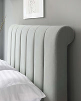 An elegant upholstered headboard with a curved silhouette and ribbed vertical panelling, in a soft grey fabric, set against a grey wall with a minimalist framed line drawing above.