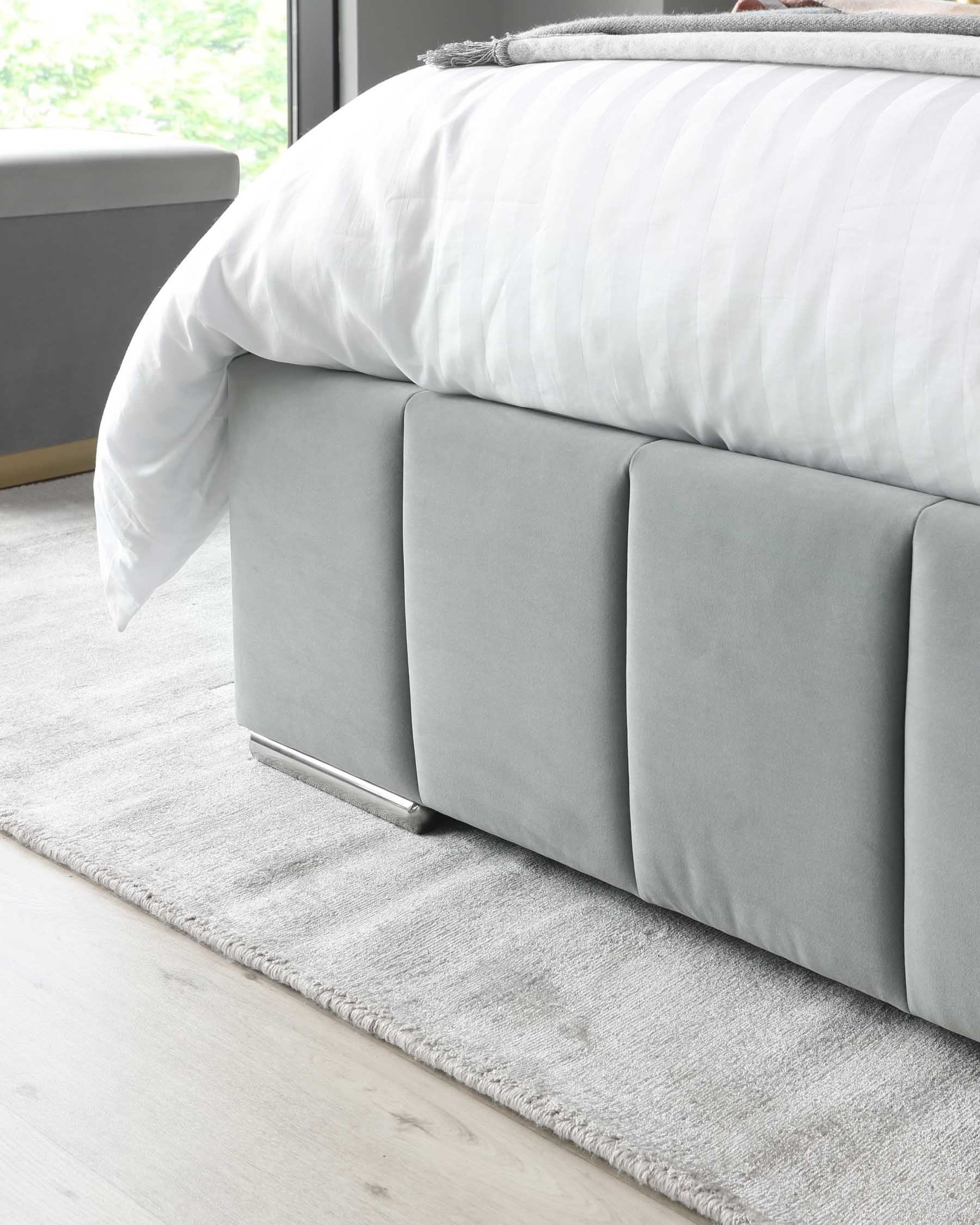 Modern minimalist light grey upholstered bed with a tufted headboard and sleek silver metal legs, topped with a white striped comforter and a grey throw on a plush grey area rug.