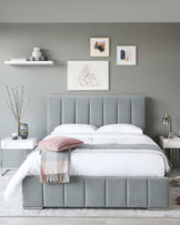 Contemporary bedroom featuring a large upholstered bed with a vertically tufted headboard in a soft grey colour. A pair of white nightstands flank the bed, each with a sleek, minimalist design. The bedding is styled with neutral tones including white bed linens accented with a grey striped pattern and a plush throw blanket in shades of pink and grey. The room's modern aesthetic is complemented by simple wall-mounted shelves displaying decorative objects and framed artwork above the bed.