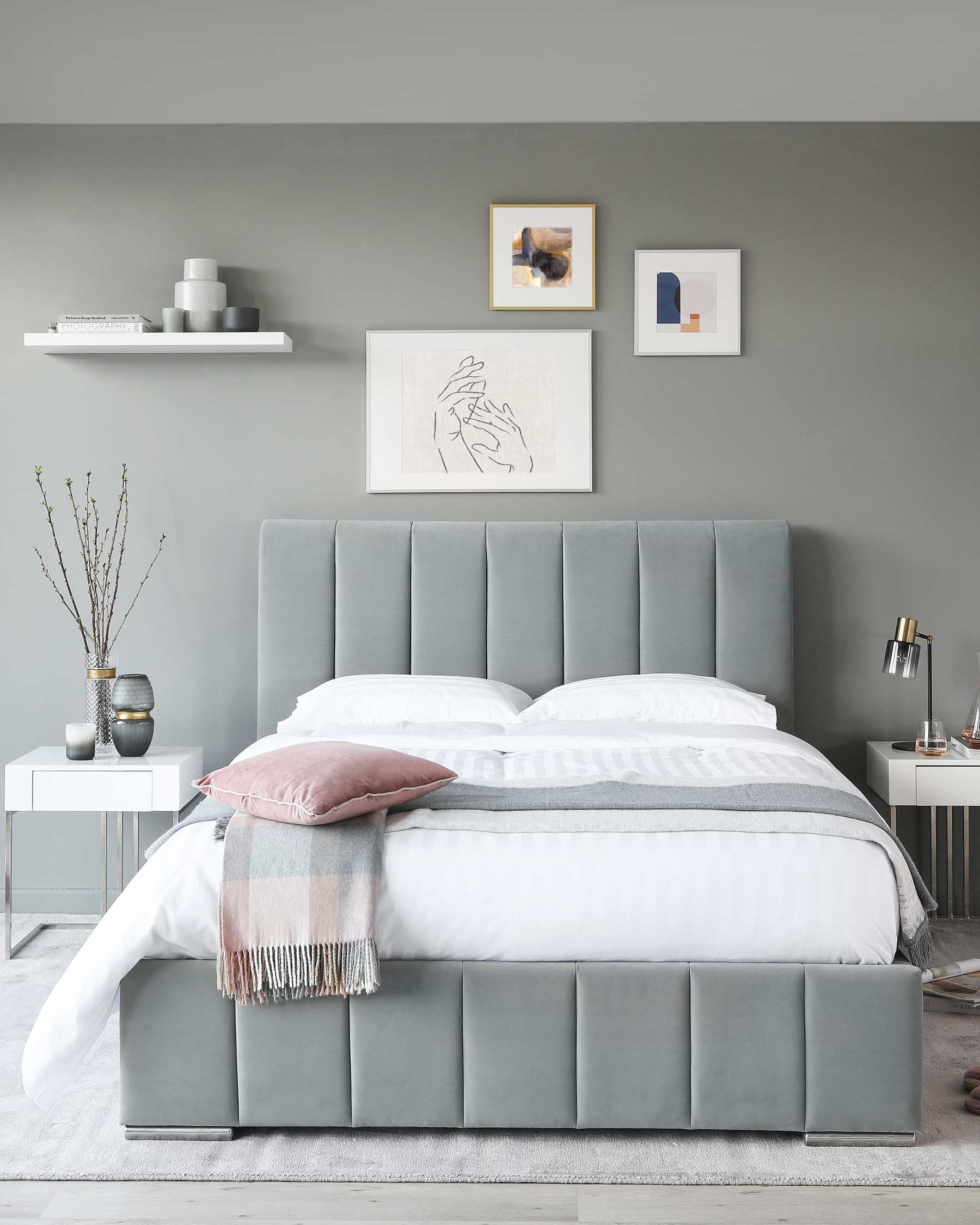 Contemporary bedroom featuring a large upholstered bed with a vertically tufted headboard in a soft grey colour. A pair of white nightstands flank the bed, each with a sleek, minimalist design. The bedding is styled with neutral tones including white bed linens accented with a grey striped pattern and a plush throw blanket in shades of pink and grey. The room's modern aesthetic is complemented by simple wall-mounted shelves displaying decorative objects and framed artwork above the bed.