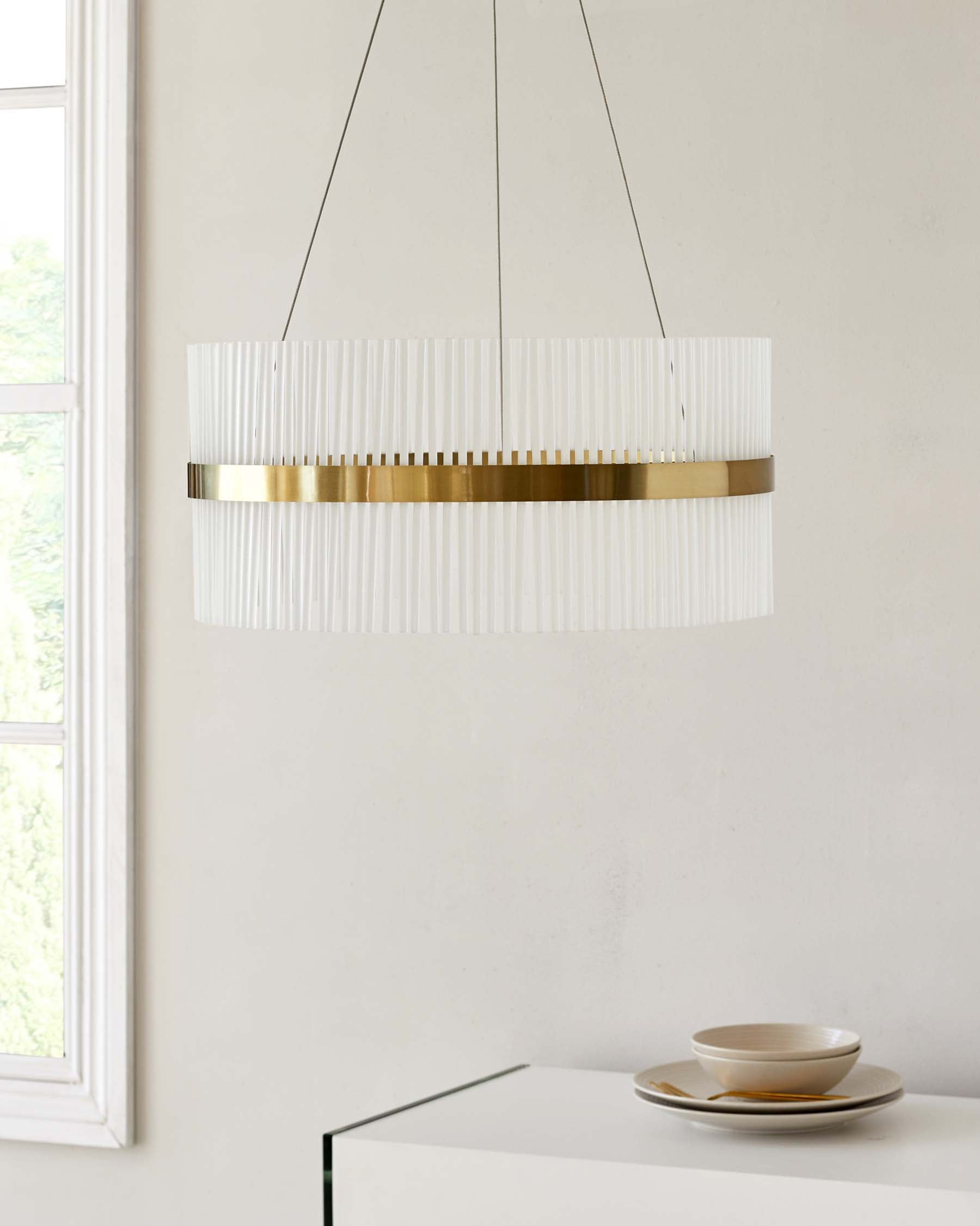 Modern minimalist white sideboard with a clean, sleek design featuring a flat top surface and seamless edges. The sideboard is partially visible, displaying a set of neutral-toned ceramic dishes on its surface. Above the sideboard hangs an elegant pendant light with a cylindrical shape, composed of vertical white slats and a lustrous gold band, emitting a warm, diffused light. The furniture and lighting fixture are set against a bright, airy room with a large window providing natural light.