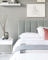 Elegant bedroom setting featuring a tall, upholstered, light grey panel headboard and a minimalist white floating bedside table. The headboard's vertical channel tufting adds a touch of sophistication, complementing the simple, clean lines of the sleek bedside table poised on two transparent support legs.