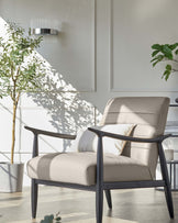 Modern armchair with a taupe leather upholstery and a minimalist black wooden frame, accompanied by a plush white cushion. The chair is in a bright, sunlit room with an indoor potted tree, giving an ambiance of natural serenity.