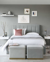 Contemporary bedroom furniture featuring a king-sized bed with a plush, grey upholstered headboard and a white bedding set. Two light grey, fabric-covered storage ottomans sit at the foot of the bed. Flanking the bed are two modern white nightstands, each adorned with decorative items.