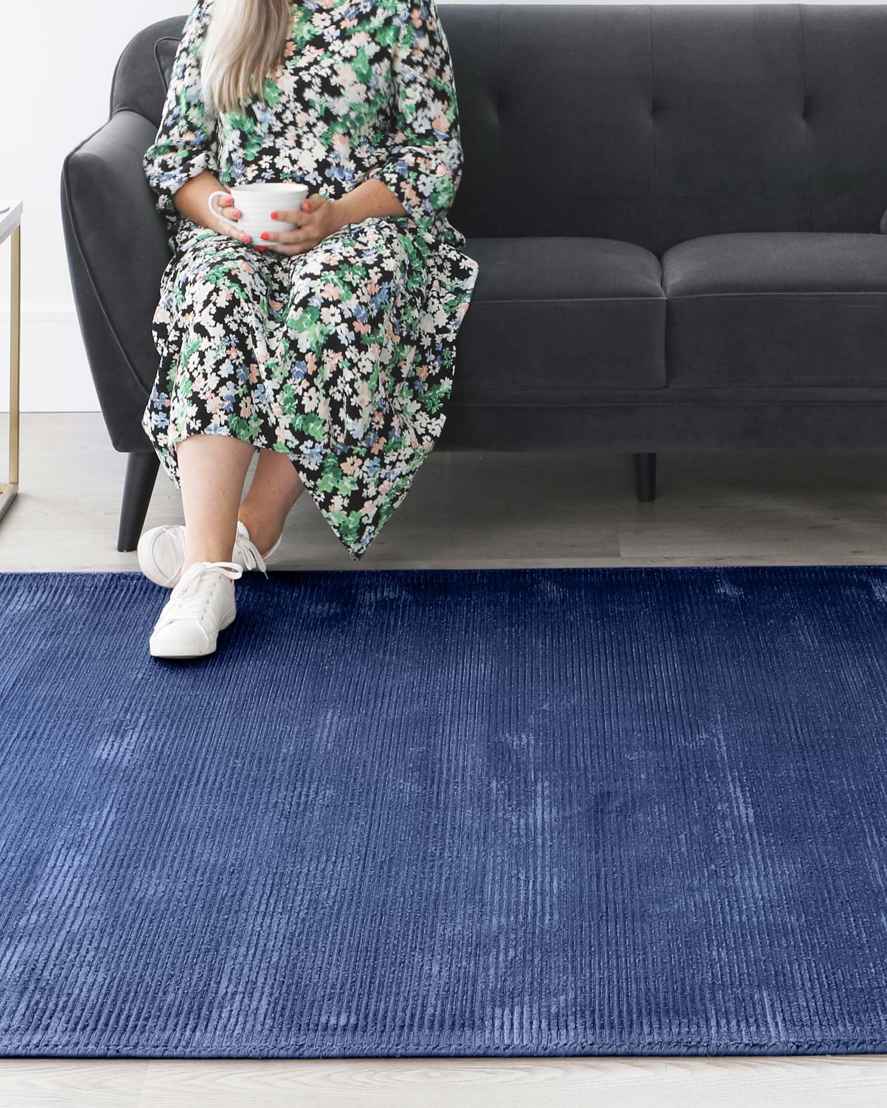 A modern three-seater sofa with a tufted, linear design in a dark grey upholstery. The sofa has cylindrical armrests and is supported by subtle, low-profile legs. A textured deep blue area rug lies on the floor in front of the sofa, adding contrast and a pop of colour to the room's decor.