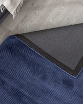 Close-up view of a textured blue area rug with a black edge, partially overlapping a grey rug on a light wooden floor, with the corner of a fringed off-white throw blanket visible in the background.