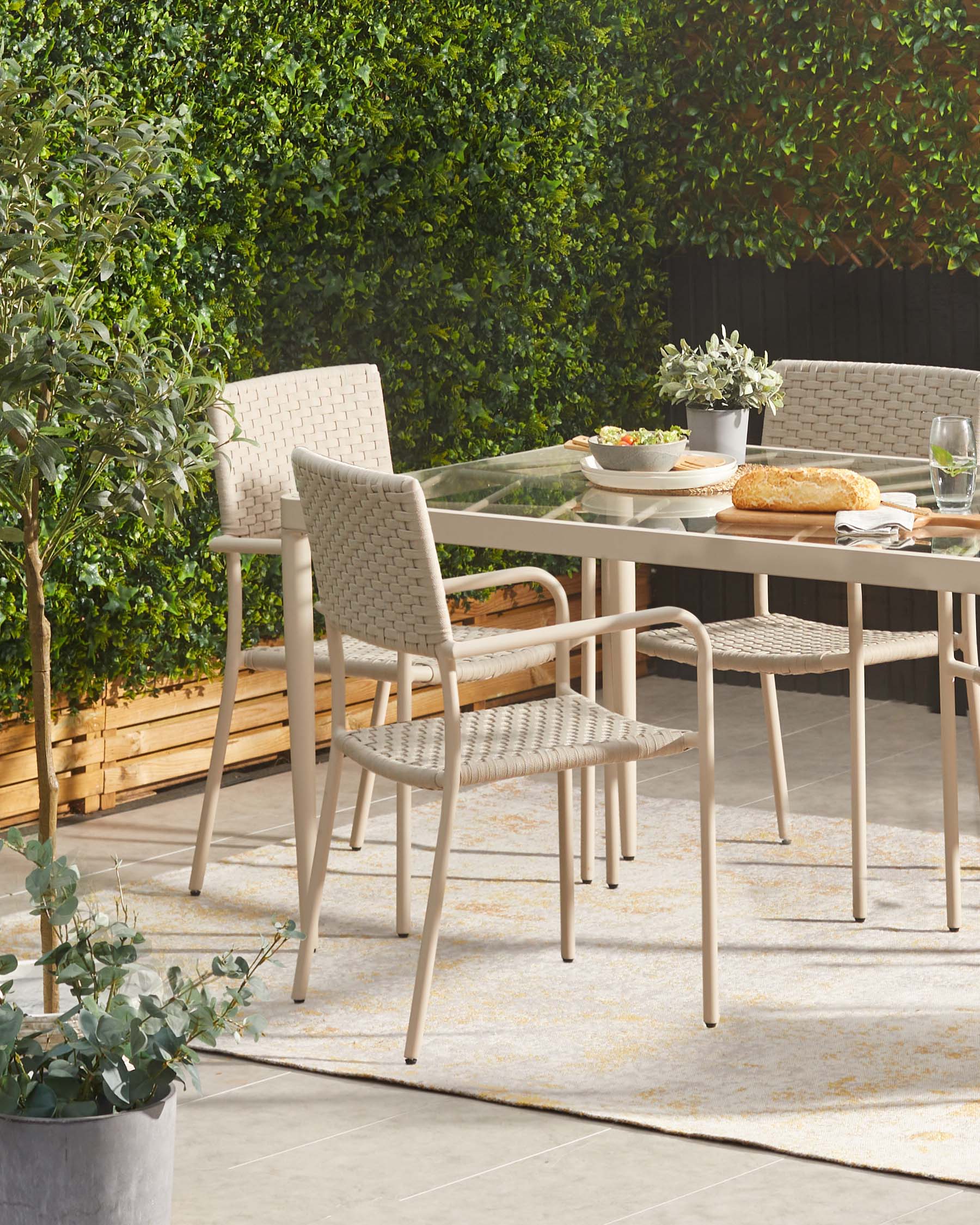 Outdoor patio furniture featuring a minimalist beige metal frame table with a glass top, complemented by matching armchairs with a woven beige back and seat design. The set is placed on a muted outdoor rug, with a background of lush greenery, showcasing a contemporary alfresco dining experience.