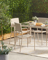 Outdoor patio set featuring a rectangular glass-top dining table with a light beige metal frame and four matching chairs with woven back and seat design on a textured beige and yellow area rug. Each chair has armrests and a gently sloping back for comfort, set against a backdrop of lush greenery. The table is accessorized with a small potted plant and a laid out casual dining setting.