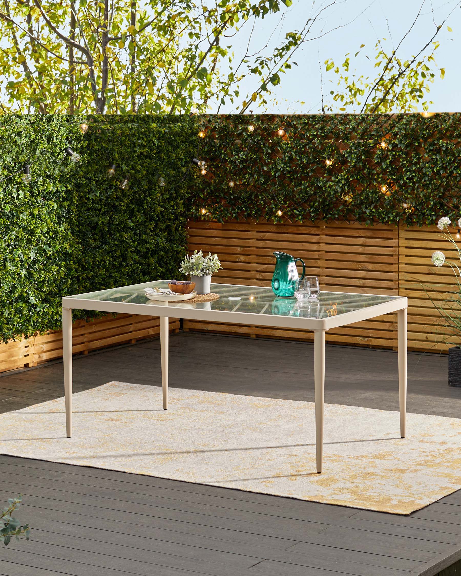 Outdoor rectangular dining table with a clear glass top and tan metal frame, featuring a modern, minimalist design, displayed on a wooden deck with a muted beige and yellow patterned outdoor rug underneath.