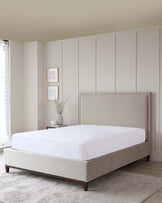 A minimalist-style bedroom featuring a contemporarily designed bed with a clean-lined, upholstered headboard in a soft beige colour. The bed has a complementary upholstered base, supported by subtle wooden legs, and is dressed in plain white bedding. To the side of the bed, there is a matching nightstand with a simple ceramic vase holding dried botanicals, contributing to the room's serene ambiance.