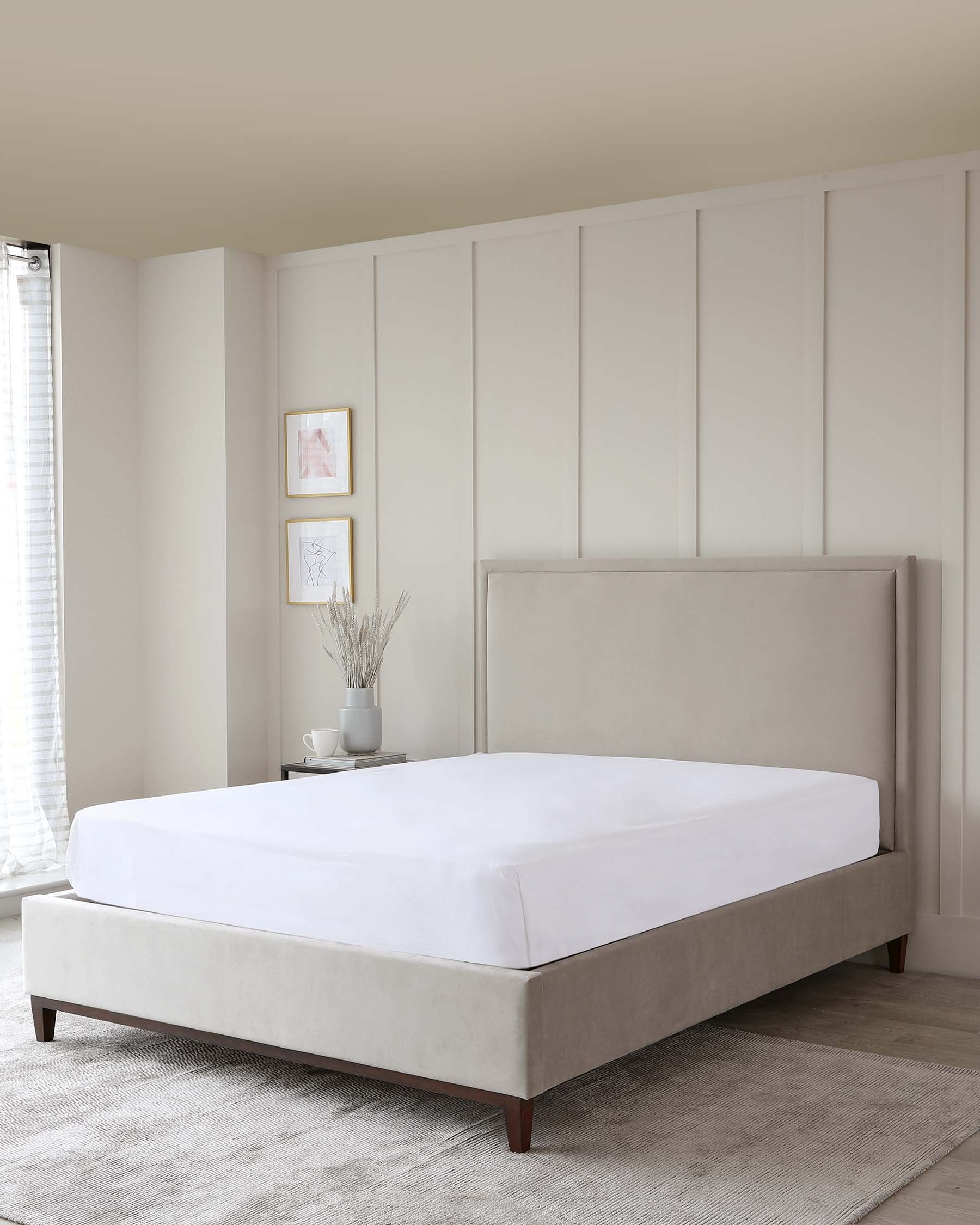 Modern minimalist upholstered platform bed with a clean-lined, padded headboard and solid wood legs, displayed in a neutrally-toned bedroom setting. A sleek bedside table with a decorative vase and two framed artworks adorns the space.