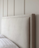 Elegant, minimalist upholstered headboard in a light beige fabric, set against a pale wall with vertical panels, partially shown with a bed dressed in a white and grey checkered duvet.