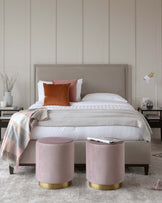 Elegant bedroom furniture featuring a large upholstered bed with a high headboard in a soft beige, flanked by two matching nightstands with a minimalist design. In front of the bed are two round blush pink ottomans with gold metallic bases, sitting on a textured light grey area rug. The bedding is crisp white with a striped pattern, accented by a throw in shades of pink and grey, and a single terra cotta decorative pillow adds a pop of colour.