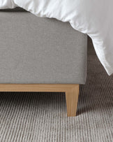 A modern minimalist bed platform with a light grey upholstered headboard and a natural wooden frame, partially covered by a crisp white duvet, set against a textured grey carpet.