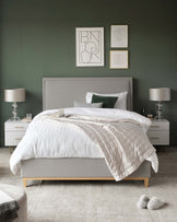 A modern bedroom setting showcasing a queen-sized bed with a minimalist grey upholstered headboard, flanked by two white and wood bedside tables with gold handles. The tables are accessorized with silver table lamps featuring white lampshades. A plush off-white area rug is partially visible at the foot of the bed.