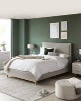 Modern bedroom furniture set featuring an upholstered platform bed with a large headboard, two white and wood nightstands, and a round, light grey ottoman at the foot of the bed. The bed is dressed with white bedding and assorted grey and green pillows. A large area rug in a light grey tone covers the floor beneath the furniture set.