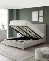 A modern light grey upholstered storage bed with its mattress lifted to reveal a spacious interior compartment, set on a muted grey rug. To the left, a coordinating white bedside dresser with a minimalist design complements the bed.