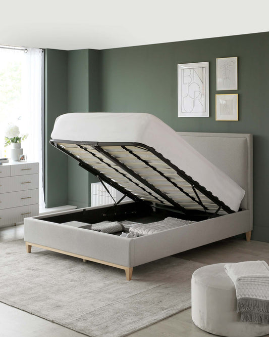 A modern light grey upholstered storage bed with its mattress lifted to reveal a spacious interior compartment, set on a muted grey rug. To the left, a coordinating white bedside dresser with a minimalist design complements the bed.