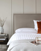 Modern bedroom setting featuring a sizeable upholstered bed headboard in a muted grey tone, coordinated with crisp white and striped bedding. Beside the bed, an elegant black nightstand with gold accents holds decorative items, including a white vase with dried botanicals.