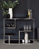 Elegant contemporary shelving unit with a semi-circular design featuring both open and enclosed shelves. The unit has reflective chrome or polished metal framing and black glass surfaces. Decorative items present include vases, glasses, cocktail shakers, and books.