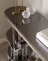 A contemporary two-tier round side table with a reflective black glass surface and polished gold metal frame, featuring a unique geometric design at each tier's junction. The table is accessorized with decorative crystal glassware and a stack of hardcover books.