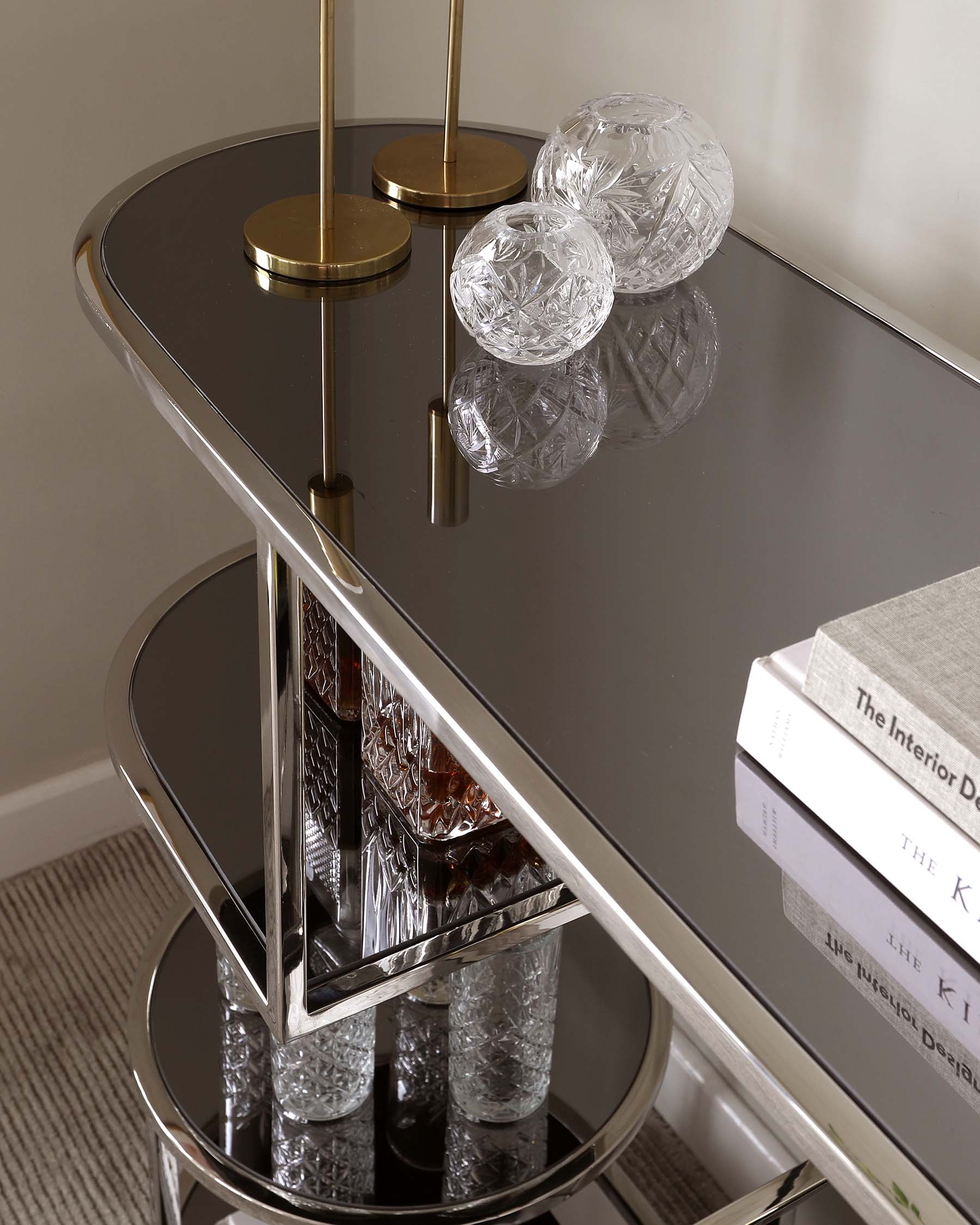 A contemporary two-tier round side table with a reflective black glass surface and polished gold metal frame, featuring a unique geometric design at each tier's junction. The table is accessorized with decorative crystal glassware and a stack of hardcover books.