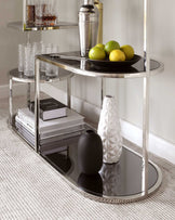 Elegant two-tiered oval bar cart with polished chrome frame and mirrored glass shelves, featuring decorative items like glassware, a cocktail shaker, and a ceramic vase on the bottom shelf, alongside a stack of books with visible titles such as "PHOTOGRAPHY." The cart is positioned on a textured, light-coloured rug against a neutral wall background.